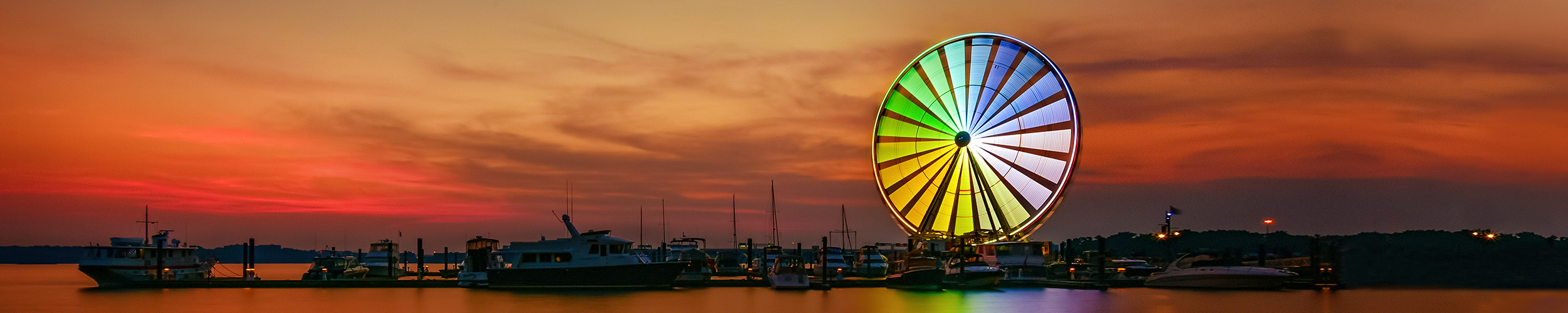 Colorful Ferris wheel glowing at sunset beside a marina filled with boats.