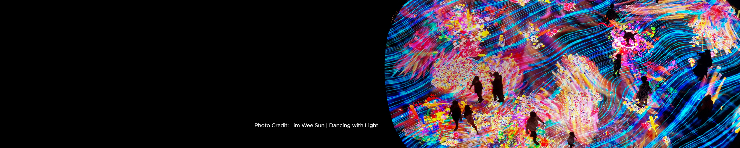 Panoramic image of an immersive digital art installation filled with vivid, swirling streams of blue, pink, yellow, and multicolored light. Silhouetted figures walk and stand across a glowing, textured floor as animated patterns flow around them, creating a dynamic, wave-like environment. Text at left reads, “Photo Credit: Lim Wee Sun | Dancing with Light.”