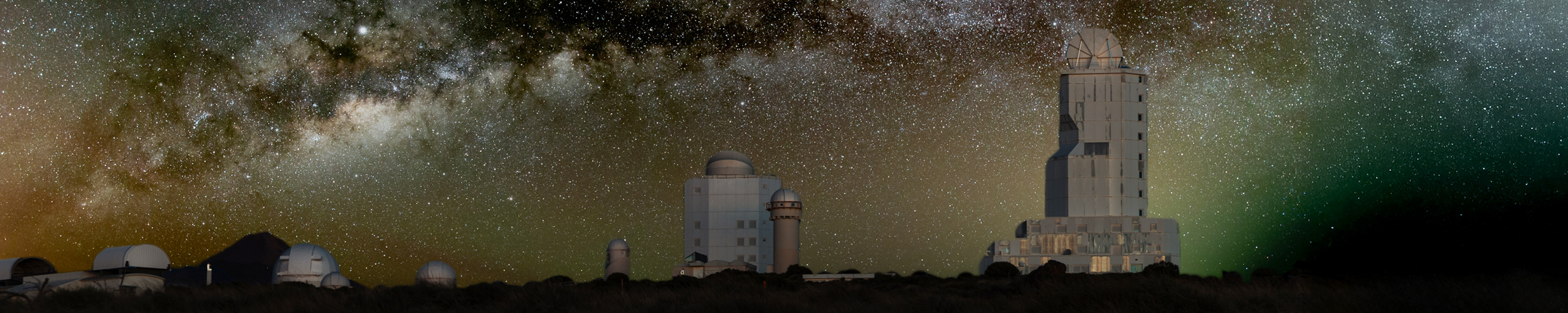 A panoramic night view of an astronomical observatory, is shown under a star‑filled sky. Multiple telescope domes and a tall, rectangular observatory tower are silhouetted against the horizon. Above them, the Milky Way stretches across the sky in a bright, detailed band of stars and dust clouds.