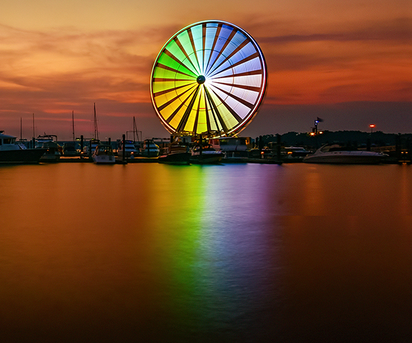 Colorful Ferris wheel glowing at sunset beside a marina filled with boats.