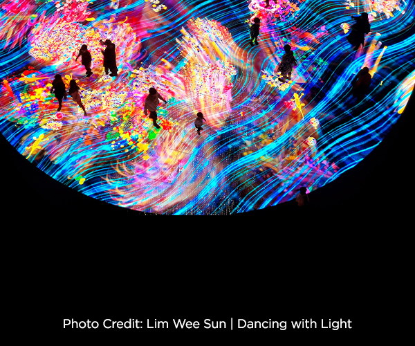 Image of an immersive digital art installation filled with vivid, swirling streams of blue, pink, yellow, and multicolored light. Silhouetted figures walk and stand across a glowing, textured floor as animated patterns flow around them, creating a dynamic, wave-like environment. Text at left reads, “Photo Credit: Lim Wee Sun | Dancing with Light.”