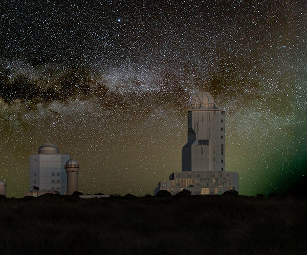 Night view of an astronomical observatory, is shown under a star‑filled sky. Multiple telescope domes and a tall, rectangular observatory tower are silhouetted against the horizon. Above them, the Milky Way stretches across the sky in a bright, detailed band of stars and dust clouds.