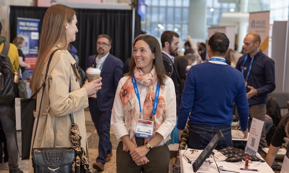 Two people standing and talking at a conference booth area, one holding a coffee cup and the other wearing a floral scarf with a conference badge. Several other attendees are visible in the background near tables with promotional materials and signage.