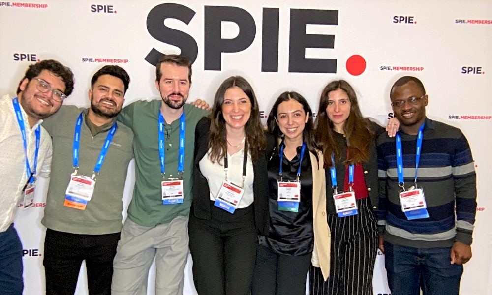 opilotHere’s a clear and modern alt text for the image: "Seven individuals wearing conference badges stand in front of a SPIE-branded backdrop at a professional event, indicating participation in an optics and photonics conference.