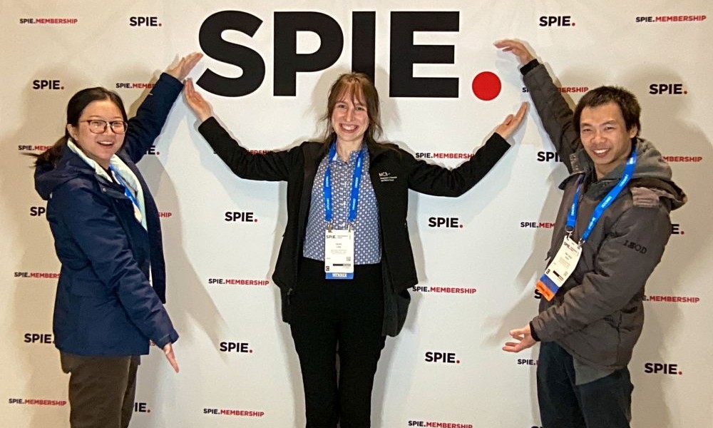 Three SPIE Student Members pose with their arms extended in different directions to frame the SPIE photo booth backdrop