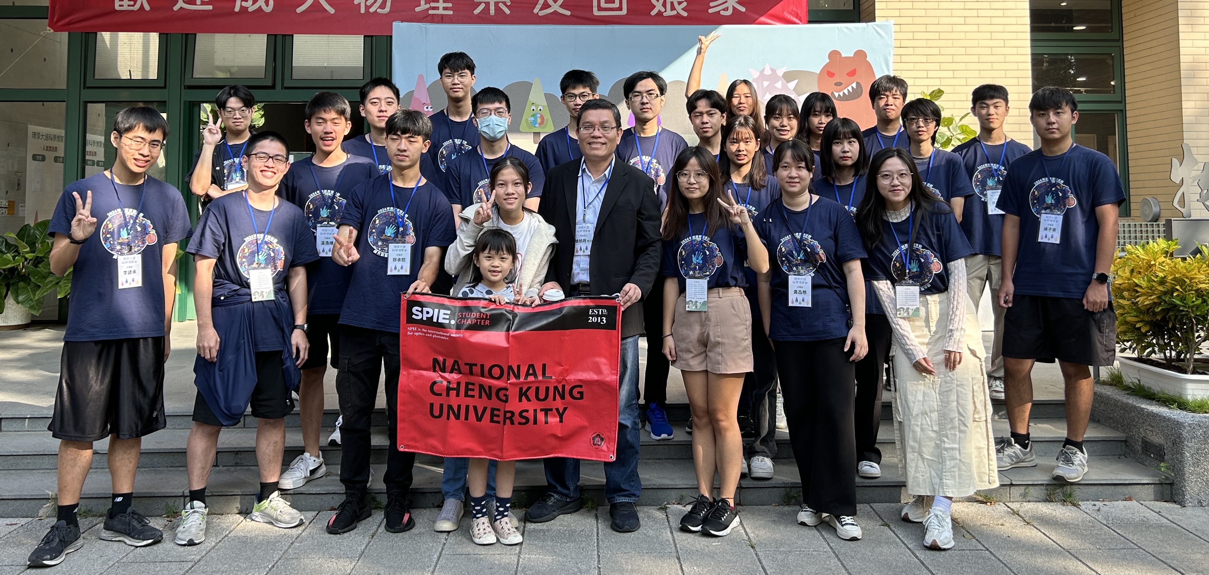 A group of students of the National Cheng Kung University SPIE Student Chapter wears matching blue t-shirts with circular designs on the front and stands in front of a building. They are holding a red banner that reads ‘SPIE Student Chapter - National Cheng Kung University.’ 