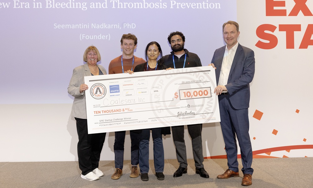 Five people stand on a stage holding an oversized $10,000 award check labeled for Coalesenz, Inc., during the 2026 SPIE Startup Challenge. A large presentation screen behind them references advances in bleeding and thrombosis prevention, and the group poses together in business attire at the event.