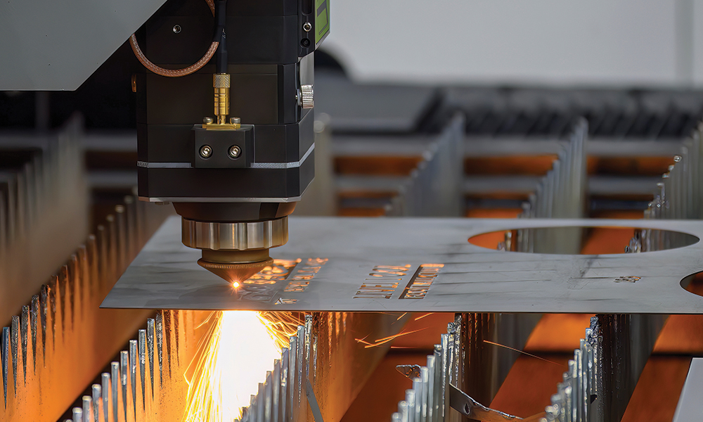An industrial laser cutting machine directs a focused beam onto a flat sheet of metal, producing bright sparks as it cuts precise shapes. The metal sheet rests on a slatted support bed inside a manufacturing setting.