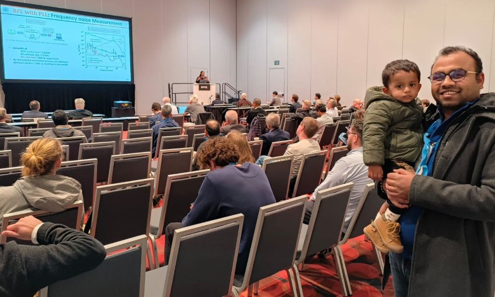 Roopa Prakash presents her research at a Photonics West 2026 conference session, while her partner stands at the back of the room holding their young child among seated attendees.
