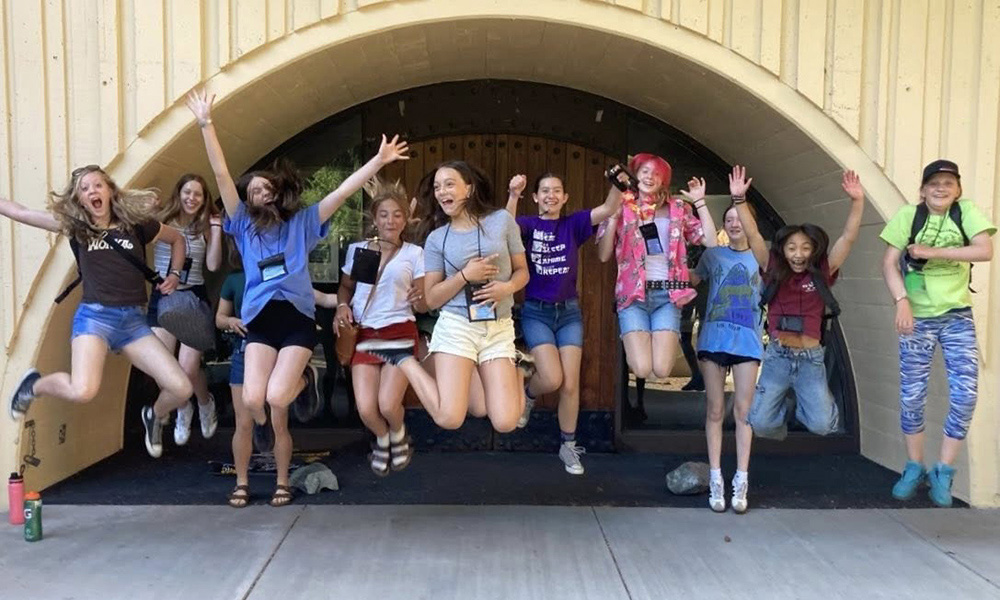 A group of ten middle school girls jump for joy in front of an archway
