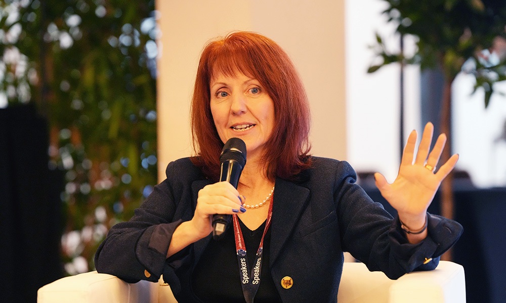 Conference speaker seated on a stage chair, holding a microphone and raising one hand, wearing a dark blazer and lanyard, with plants in the background.