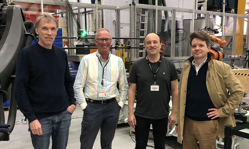 Four adults wearing visitor badges stand side by side on a factory or laboratory floor, with large industrial machinery, metal structures, and safety barriers visible behind them.