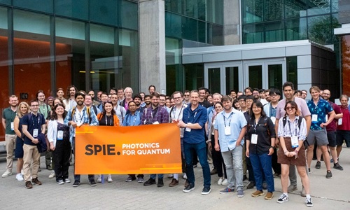 A few dozen people stand together holding an orange sign that says SPIE Photonics for Quantum