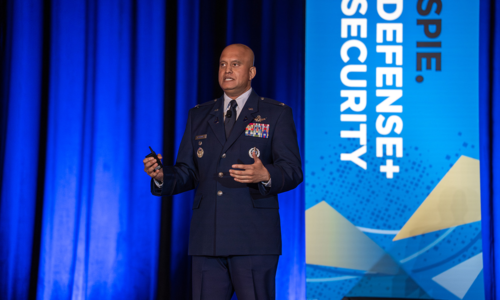 A person in a military uniform stands on a stage delivering a presentation, holding a remote clicker, with a blue curtain backdrop. A vertical banner to the right reads &ldquo;SPIE Defense + Security,&rdquo; indicating the event and conference setting