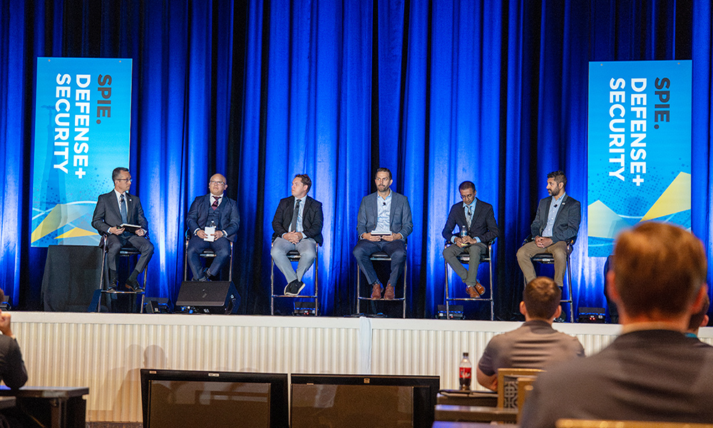 Wide shot of conference stage shows six panelists seated on stools in front of blue curtains at a professional event, with two vertical banners reading “SPIE Defense + Security” on either side. An audience is visible in the foreground.
