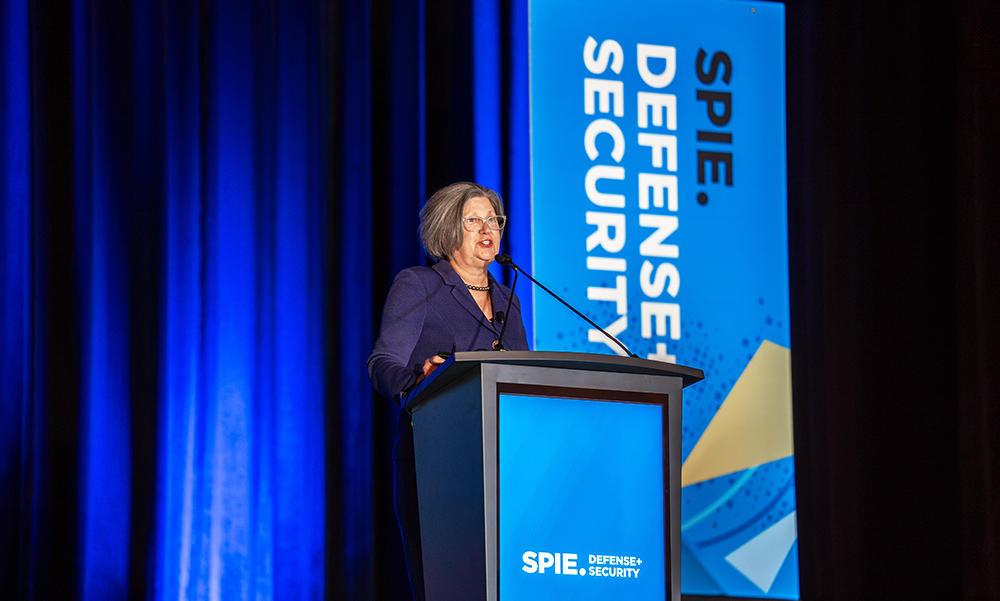 A person stands at a podium delivering a plenary talk on a stage, with blue curtains and professional stage lighting in the background. A tall banner and the podium display the text “SPIE Defense + Security,” identifying the conference setting