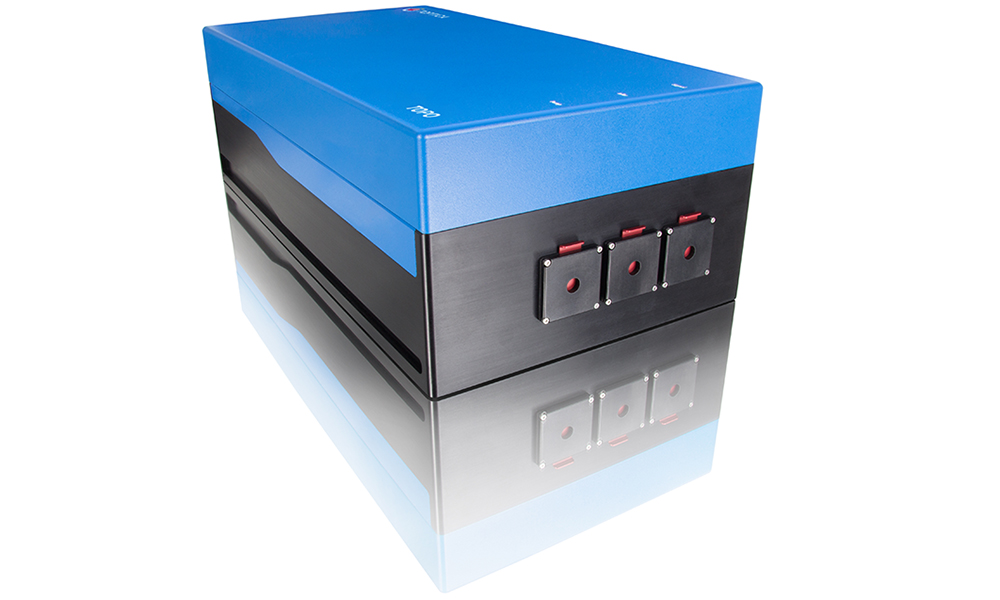 A rectangular laboratory laser instrument with a blue top and black sides sits on a reflective surface. The front panel shows three square control modules with small red indicator lights and toggle switches, suggesting a precision tunable laser system.